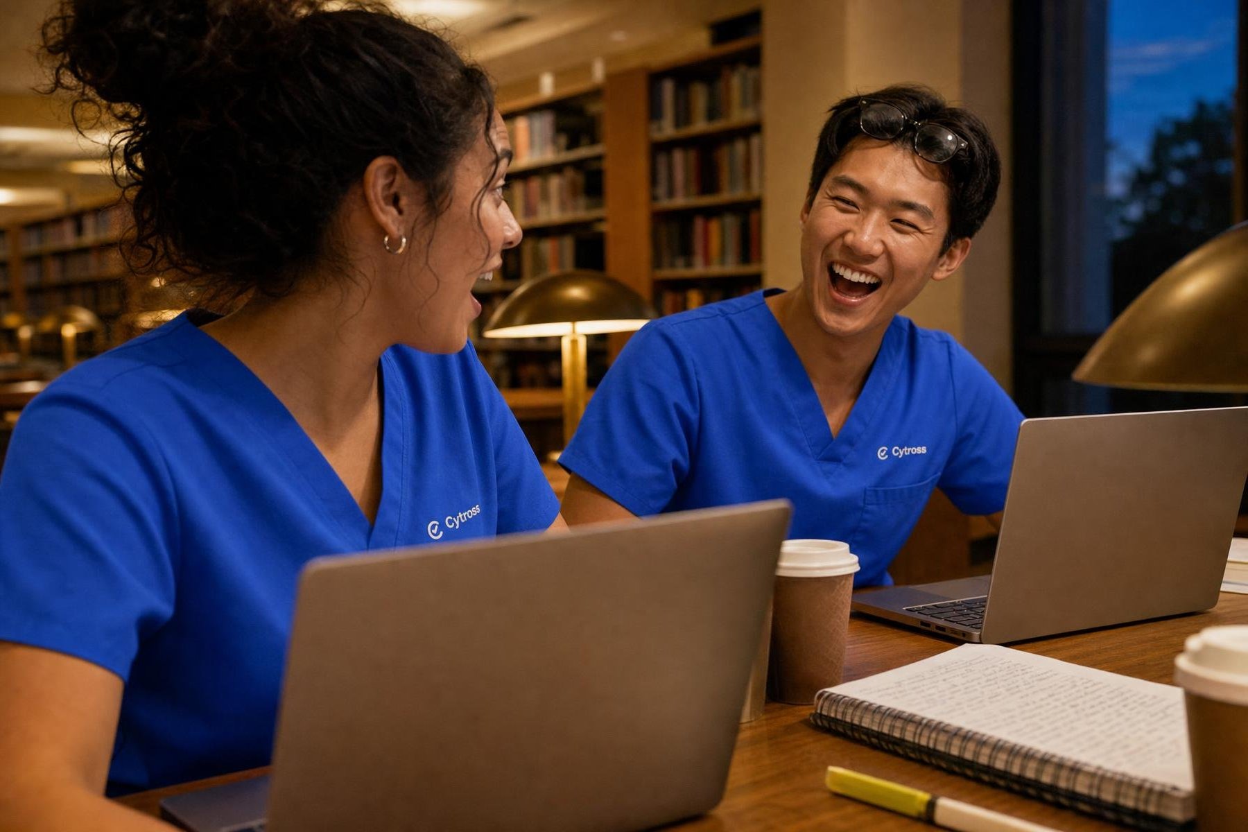 Two nursing students in royal-blue Cytross Nursing scrubs at a library table in the evening, laughing over their laptops mid-Duel.